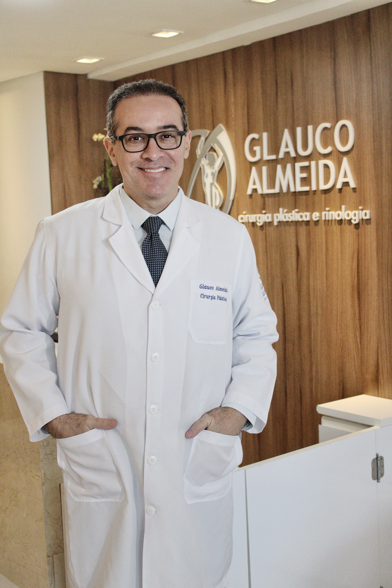 Dr. Glauco Almeida de jaleco na recepção do consultório, com a marca ao fundo